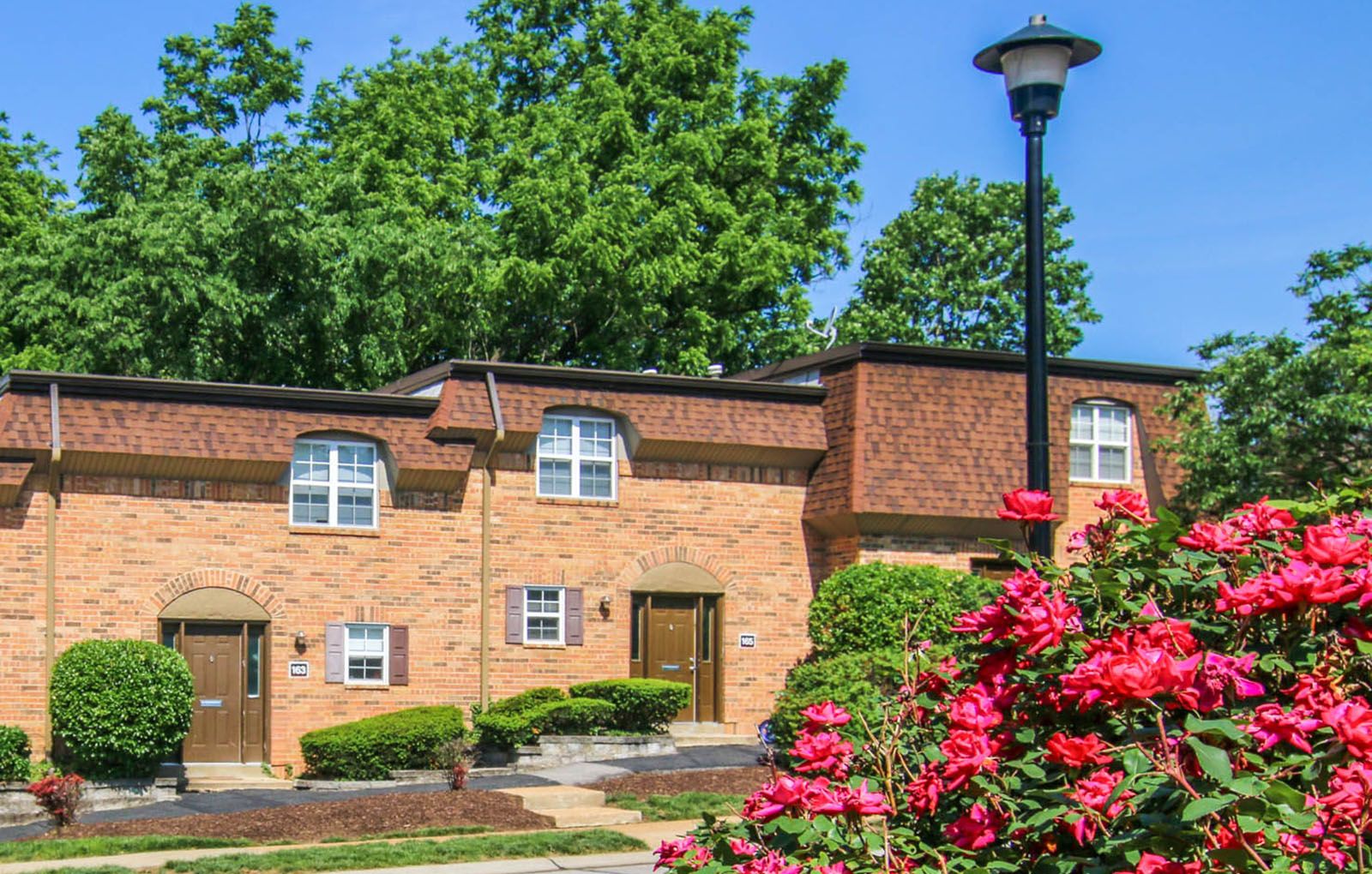 Rockwood Apartments Two brick townhouses with trimmed bushes, a lamppost, and blooming pink flowers in the foreground.