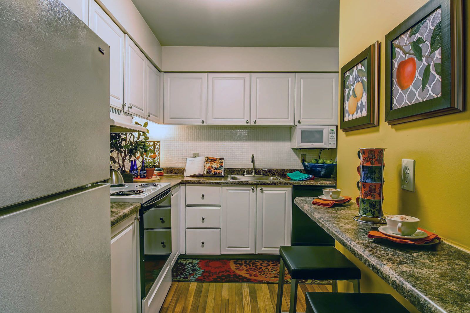 Rockwood Apartments Small kitchen with white cabinets, a fridge, stove, bar counter, and colorful fruit artwork on a yellow wall.