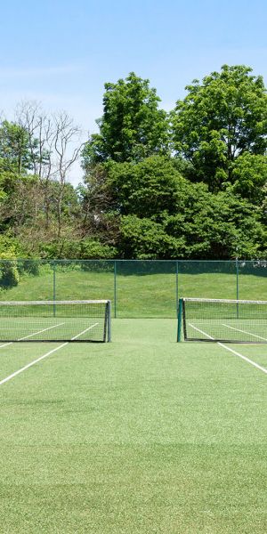 Rockwood Apartments Empty grass tennis court surrounded by trees and greenery on a sunny day.