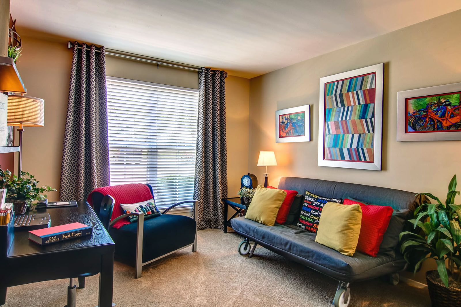 Rockwood Apartments Cozy home office with a futon, pillows, armchair, desk, plants, and colorful wall art near a window.