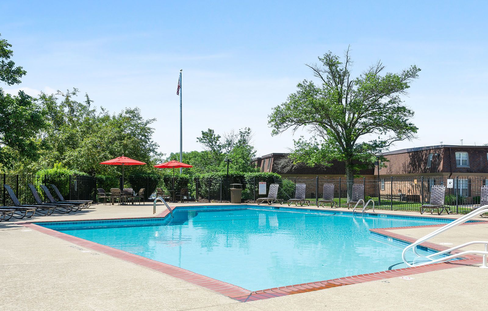 Rockwood Apartments Outdoor swimming pool with lounge chairs, red umbrellas, and trees under a clear blue sky.