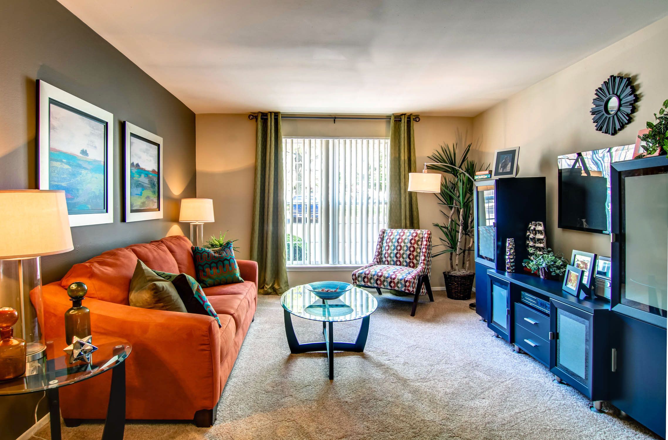 Rockwood Apartments Colorful living room with an orange sofa, patterned chair, TV, glass table, and large window with curtains.