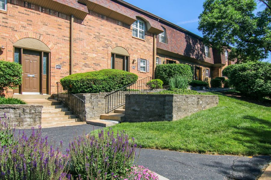 Rockwood Apartments Brick townhouse with brown doors, trimmed bushes, stairs, and green lawn on a sunny day.