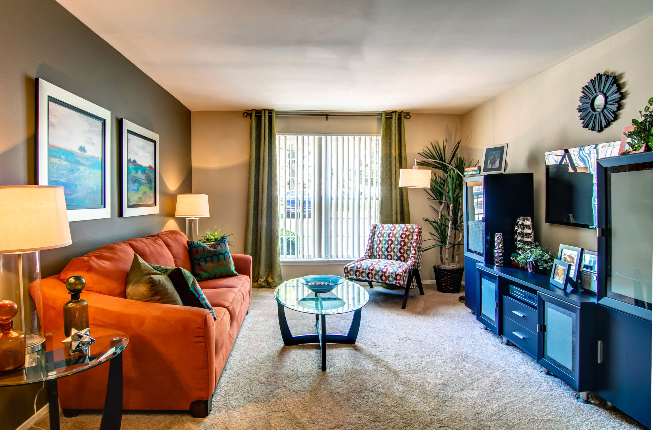 Rockwood Apartments Colorful living room with an orange sofa, patterned chair, TV, glass table, and large window with curtains.